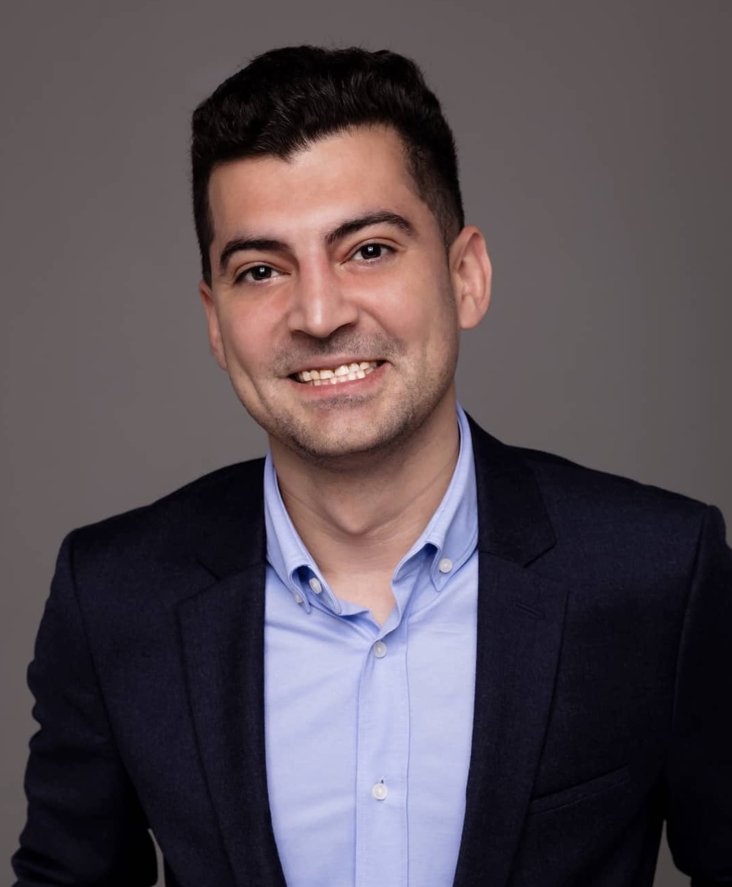 Portrait professionnel d'un membre de l'équipe souriant en costume noir et chemise bleue sur fond gris